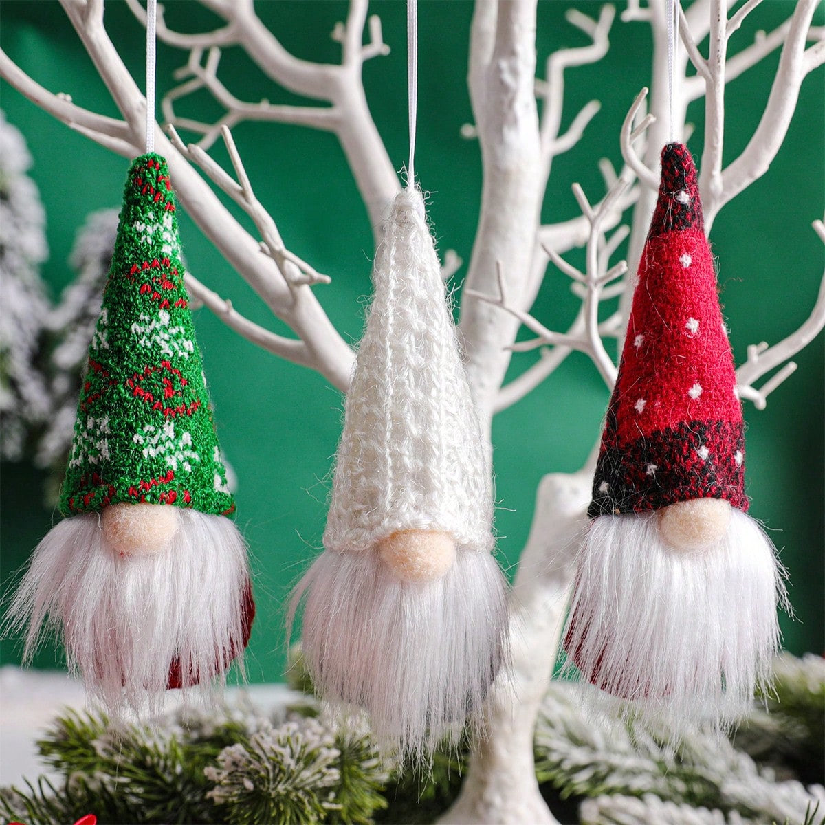 Three knitted gnome ornaments with colorful hats hanging on a branch against a green background.